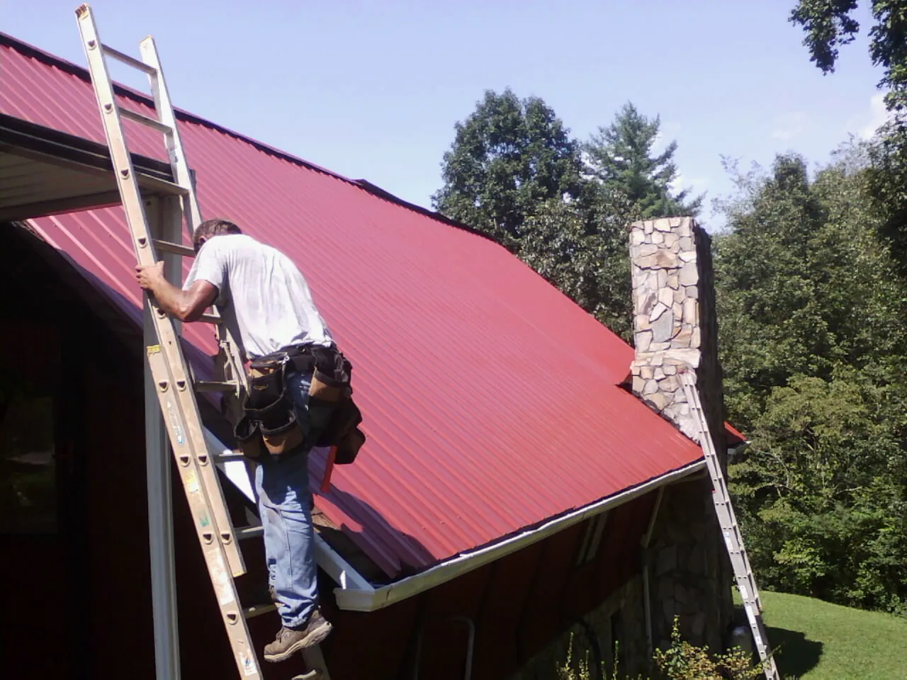 Roofer on a ladder beside a red metal roof and stone chimney — Soffit & Fascia Repair by Roofing Brothers in Fort Sill